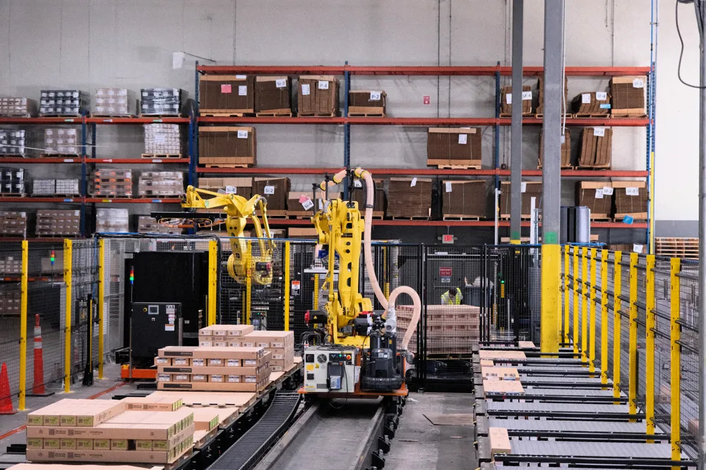 Two yellow robot arms stacking cases onto pallets inside a fenced cell in a warehouse.